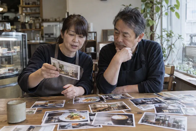 カフェの店主夫婦が、テーブルに並べた料理の写真から載せるものを選んでいる