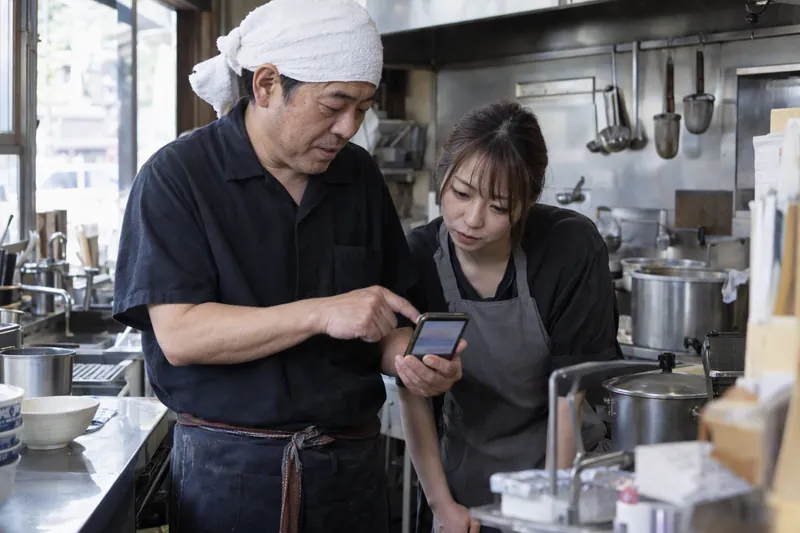 麺の店の厨房の脇で、店主がスマートフォンの画面を指さして家族と直したい所を話している