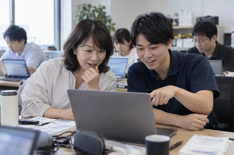 2人の社員さんが1台のノートパソコンに顔を寄せ、出てきた結果を指さして見ている