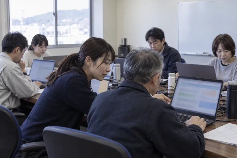会議室で社員さんがそれぞれノートパソコンを開き、隣の人の画面をのぞいて動いたかを確かめている